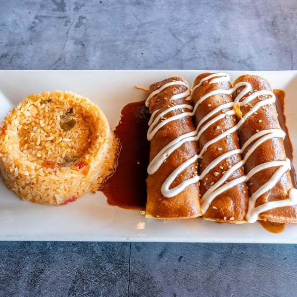 Three enchiladas with mole sauce, crema, and sesame seeds, served with a side of Mexican rice.